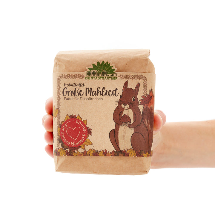 Hand hält eine Packung Eichhörnchenfutter mit der Aufschrift "Große Mahlzeit" und einem Bild eines Eichhörnchens darauf.