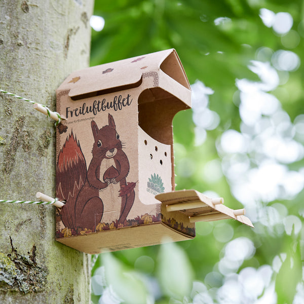Vogelhaus aus Karton mit Eichhörnchen-Aufdruck an einem Baum befestigt, beschriftet mit "Freiluftbuffet" in einem grünen Garten.