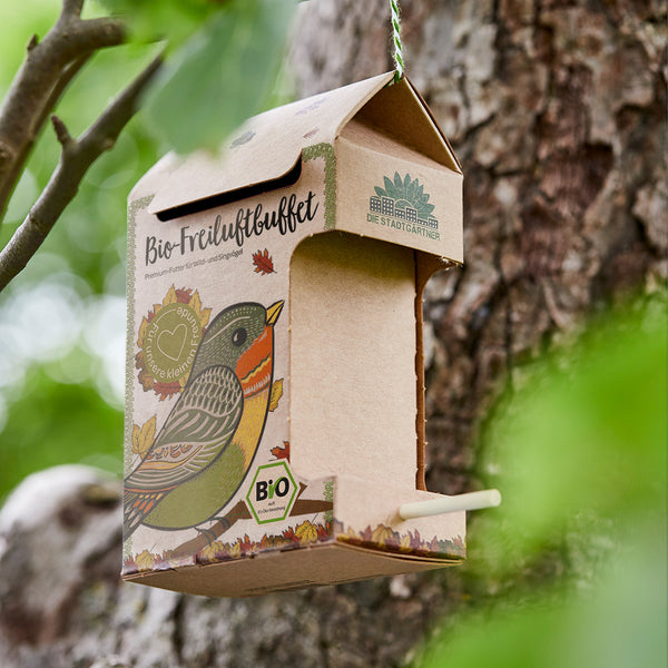 Bio-Vogelfutterhaus an einem Baum befestigt mit Illustrationen von Vögeln und Blättern auf der Verpackung.