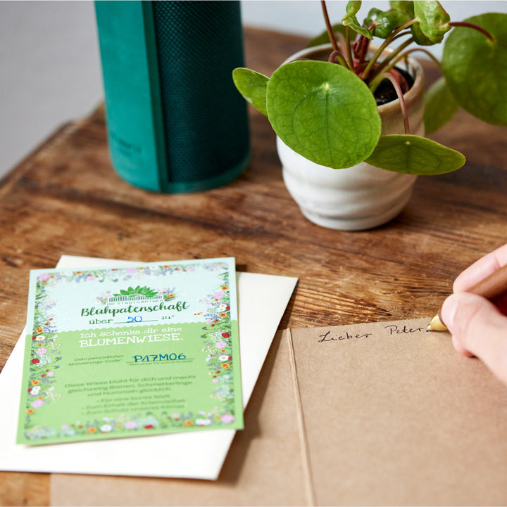 Eine Hand schreibt auf einem Umschlag neben einer Geschenkkarte über Blumenpatenschaft und einer Pflanze auf einem Holztisch"