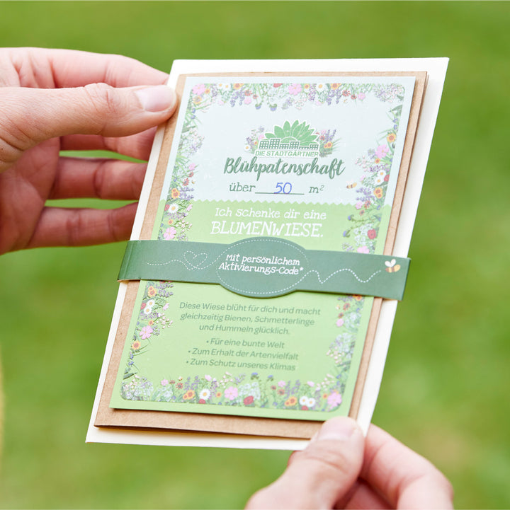 Hände halten eine Geschenkkarte für eine Blühpeterschaft über 50 Quadratmeter mit blumigem Design und Text darauf.