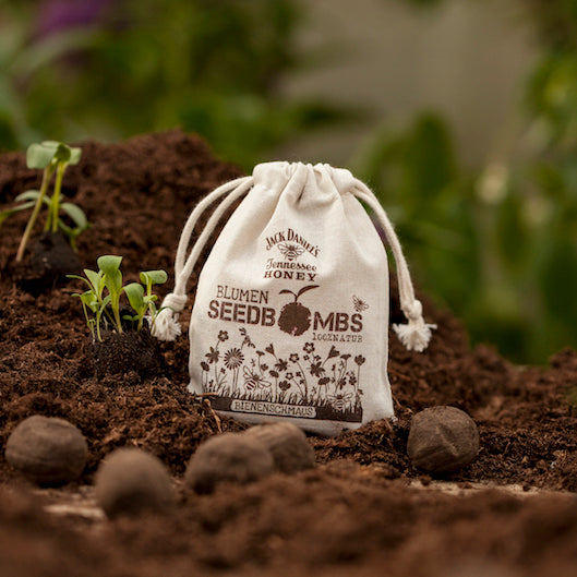 Ein kleiner Stoffbeutel mit der Aufschrift "Blumen Seedbombs" steht auf fruchtbarer Erde neben Keimlingen und Samenbomben.