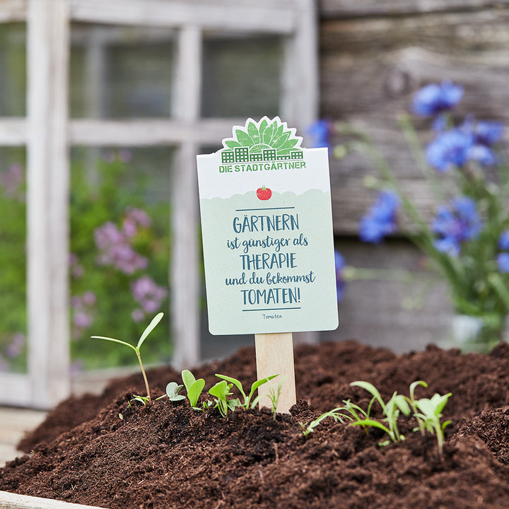 Gartensteckschild mit der Aufschrift "Gärtnern ist günstiger als Therapie und du bekommst TOMATEN!", umgeben von Erde und jungen Pflanzen.
