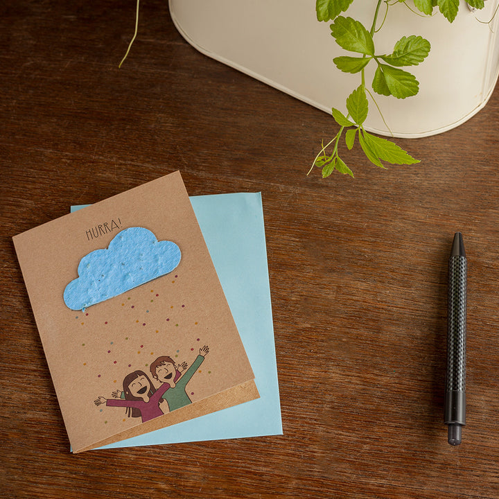 Glückwunschkarte mit jubelnden Personen und einer Wolke auf einem Tisch neben einem Stift und Pflanzenblättern.